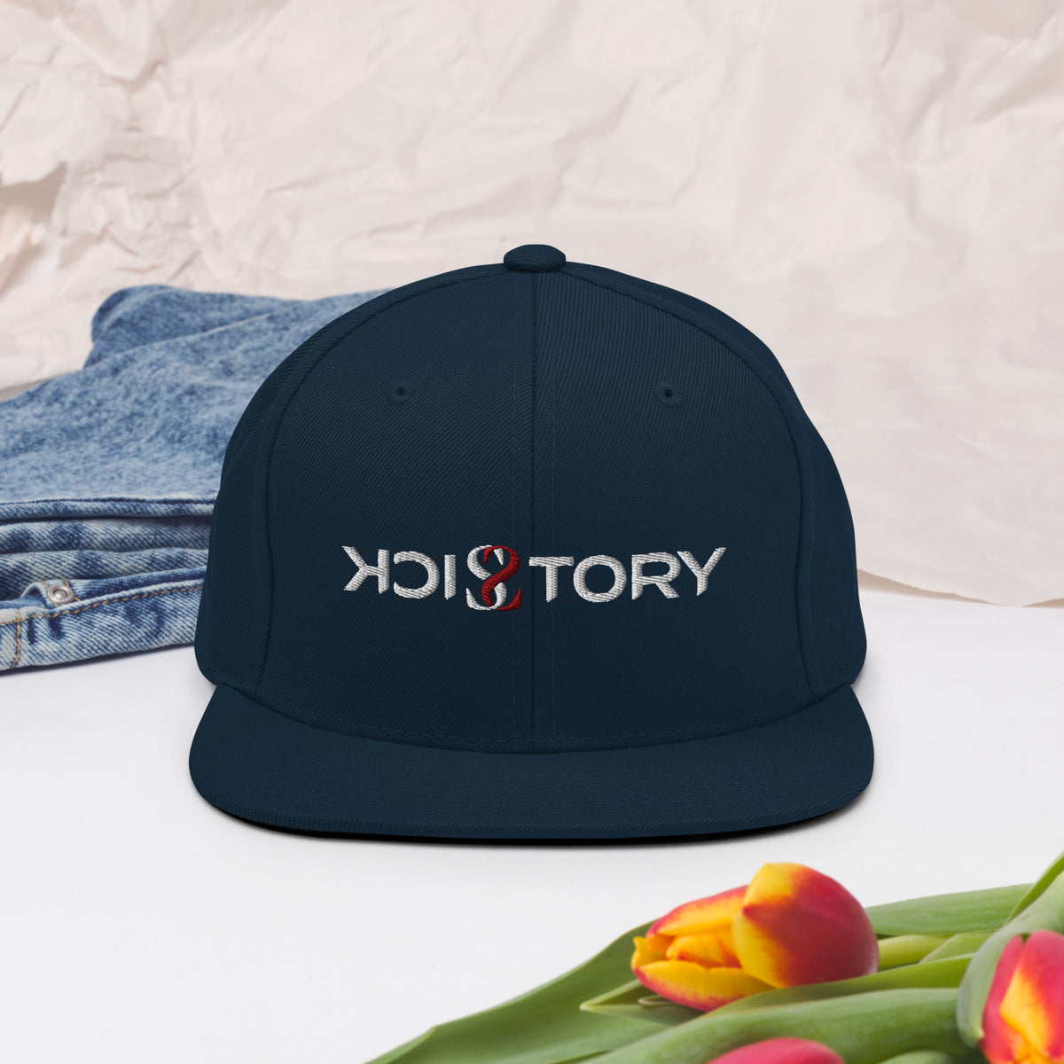 Dunkelblaue Sick Story Snapback Cap mit besticktem Logo und flachem Schirm neben einer Jeans und Tulpen